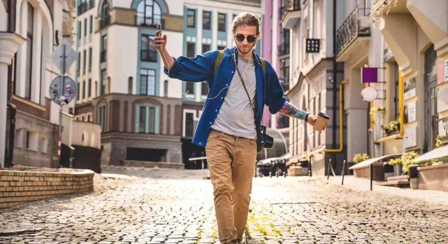Giovane con caffè e smartphone cammina in città su un selciato, godendosi la musica e il paesaggio urbano.