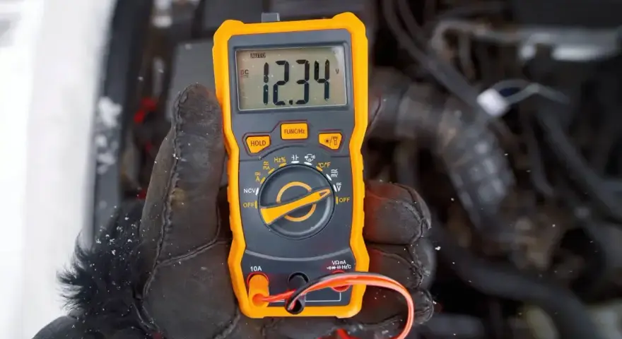 Ręka w rękawicy trzyma pomarańczowy multimetr, który pokazuje 12.34V. Auto nie odpala, akumulator sprawny, problem leży gdzie indziej.