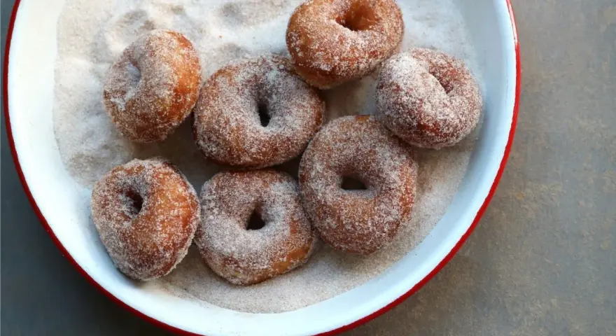 Små, nybakade donuts som rullats i socker och kanel ligger i en vit skål med röd kant.