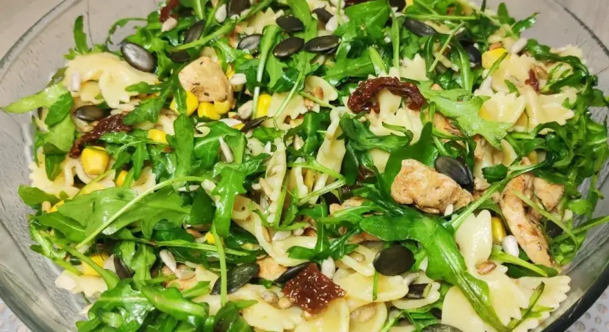 Kolorowa sałatka z makaronem farfalle, rukolą, kurczakiem, kukurydzą i suszonymi pomidorami, posypana pestkami dyni i słonecznika.