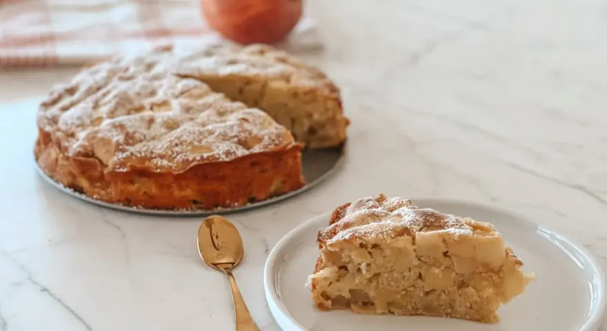 Une part de **crépiau aux pommes** saupoudrée de sucre glace, à côté d'un gâteau entier et d'une pomme. Une cuillère dorée est posée sur l'assiette.