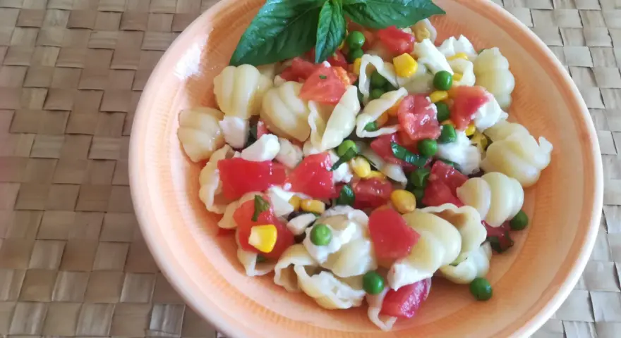 Un'appetitosa insalata di pasta vegetariana con pomodorini, piselli, mais e mozzarella, guarnita con basilico fresco.