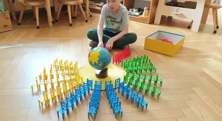 Chłopiec bawi się w przedszkolu Montessori, układając kolorowe krzesełka wokół globusa. To pokazuje, montessori co to za metoda nauczania.