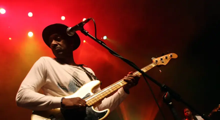 Marcus Miller w czarnym kapeluszu gra na białej gitarze basowej na tle czerwonych świateł scenicznych.