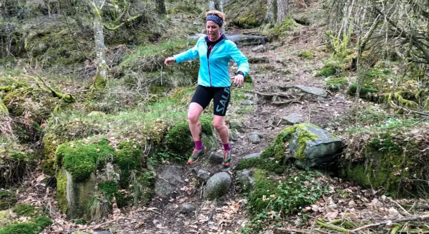 Kvinna springer i skogen, fokuserad på att springa marathon. Mossbeklädda stenar och träd skapar en utmanande terräng.