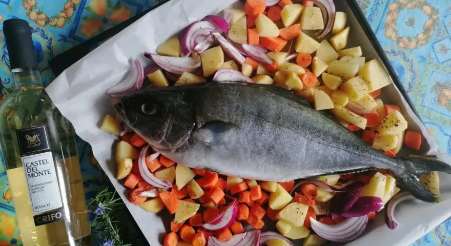 Ricciola al forno pronta per essere infornata con patate, carote e cipolle rosse. Accanto, una bottiglia di vino bianco.