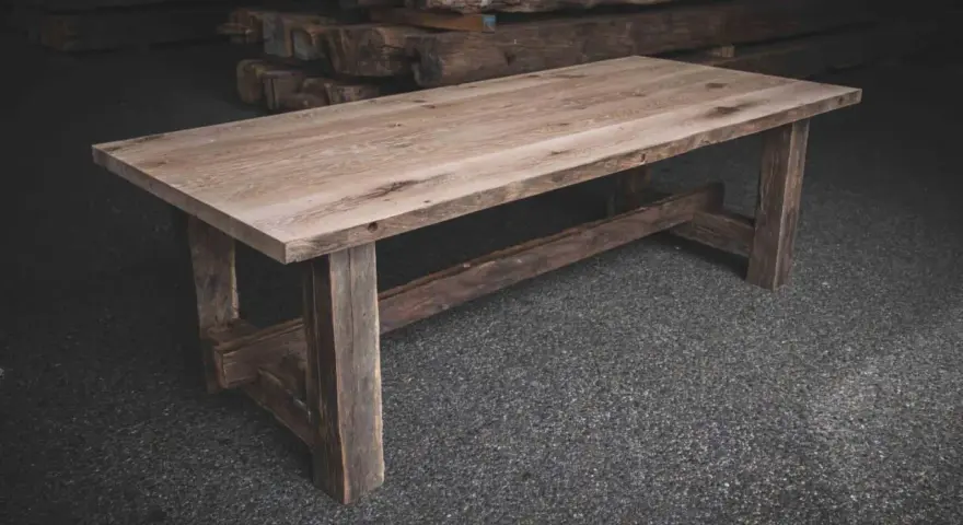 Ett rustikt bord av oljad ek med grova ben och en bred bordsskiva. Bakom syns staplade trästockar.