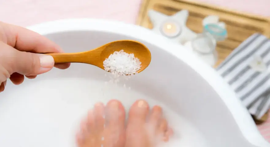 A hand adds epsom salt and baking soda to a foot soak, with feet submerged in the milky water.