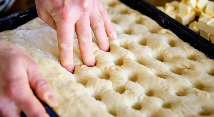 Hände drücken Vertiefungen in den Teig für ein Butterkuchen Rezept. Würfel aus Butter liegen bereit.