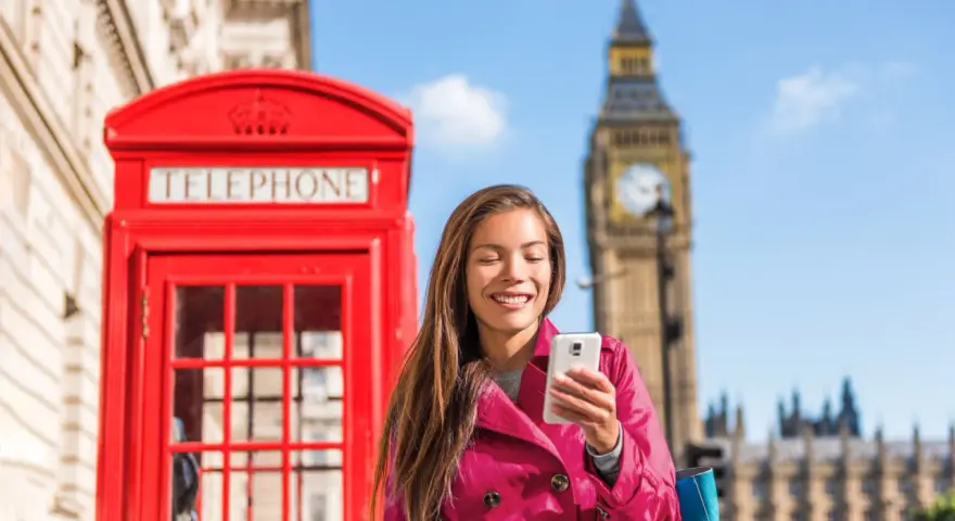 Uśmiechnięta kobieta z telefonem przed czerwoną budką telefoniczną i Big Benem. Pyta, czy w UK jest roaming, bo chce zadzwonić do domu.