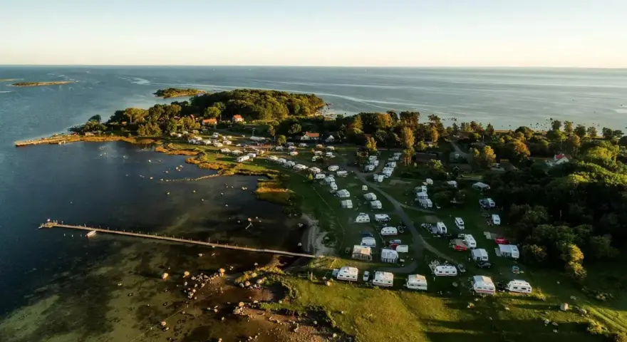 Vacker vy över Kronobergs camping med husvagnar och husbilar utspridda bland träd och vid vattnet. En brygga sträcker sig ut i det klara vattnet.