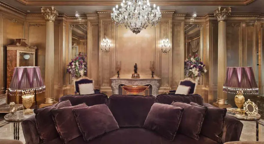 Salón lujoso con sofás de terciopelo morado, lámparas doradas y un candelabro de cristal. El techo y las paredes doradas evocan el estilo barroco español.
