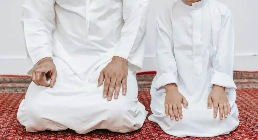 Ojciec i syn w białych strojach klęczą na dywanie, wykonując codzienną modlitwę poranną.