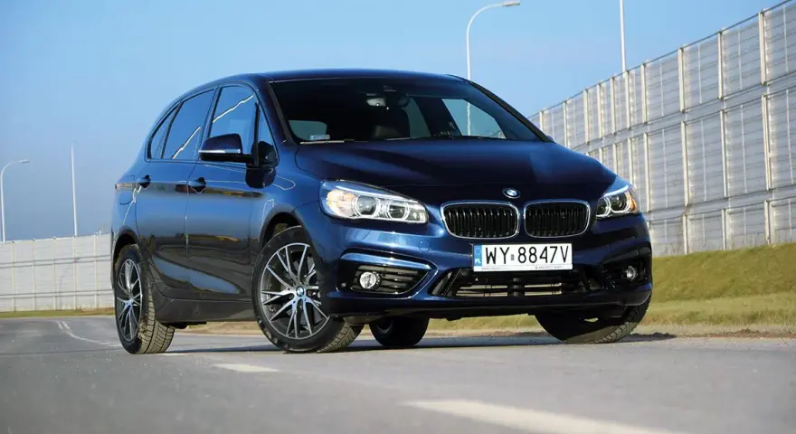 Ciemnoniebieski BMW van na tle jasnego nieba i barier dźwiękochłonnych.