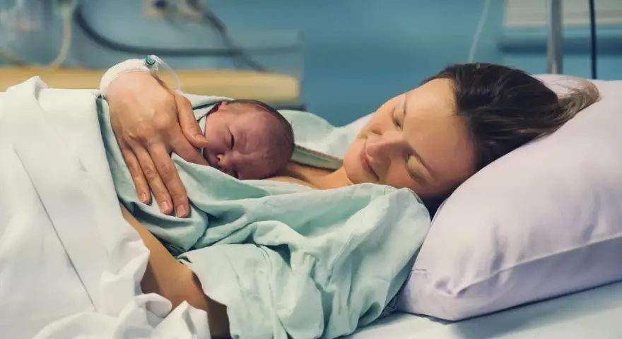 Eine Mutter hält ihr Neugeborenes im Arm, glücklich und erschöpft. Die Szene zeigt, wie lange nach Geburt im Krankenhaus die erste Bindung entsteht.