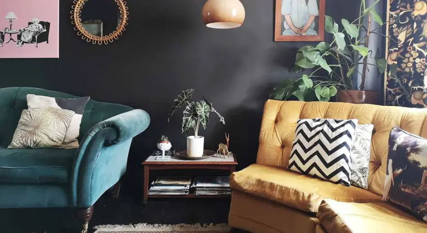 Habitación con poca luz, sofás de terciopelo verde y mostaza, cojines decorativos, plantas y arte en la pared.