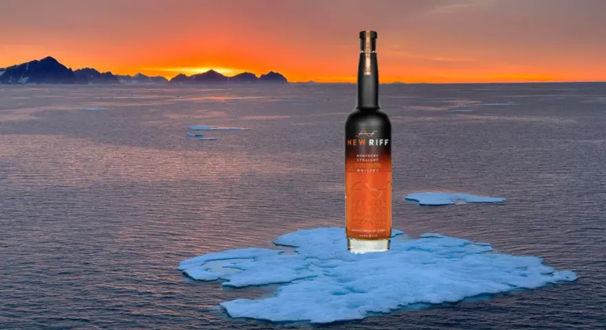 Eine Flasche New Riff Bourbon Whiskey steht auf einer Eisscholle im Meer. Der Sonnenuntergang taucht die Berge am Horizont in warmes Licht. Ein Moment der Ruhe für den besten Whisky der Welt.
