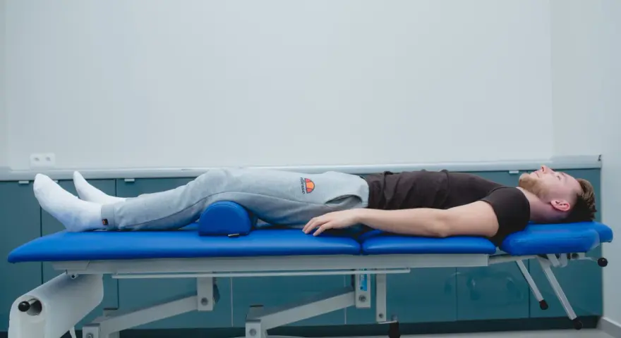 Mężczyzna leży na kozetce rehabilitacyjnej, z podparciem pod kolanami. Rozpoczęła się jego sis rehabilitacja.