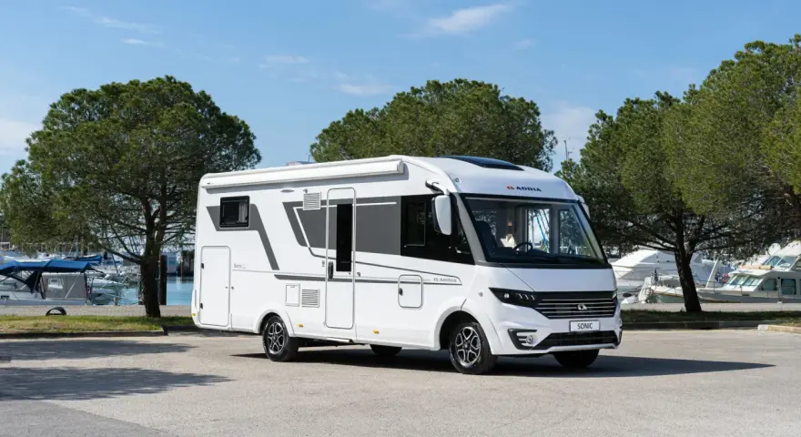 En vit Adria Sonic husbil, den lyxigaste husbilen, parkerad vid en marina med båtar och träd i bakgrunden.