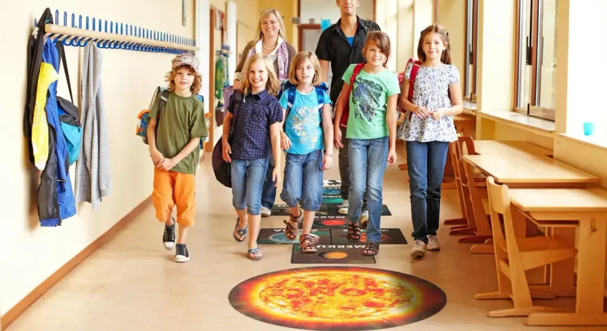 Dzieci i dorośli idą szkolnym korytarzem, stąpając po planszy z planetami. Fajne inicjatywy w szkole promują naukę przez zabawę.