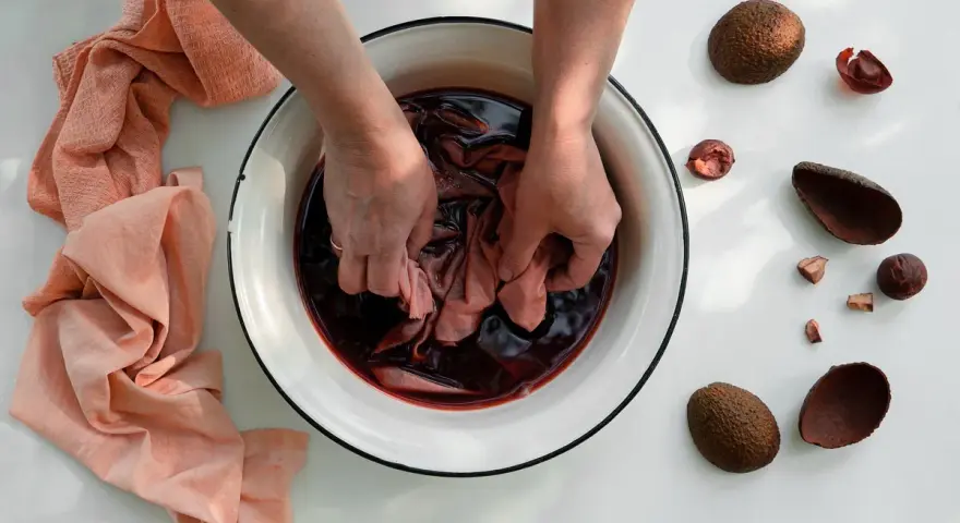 Mani che tingono un tessuto in una bacinella con un colorante naturale per tessuti. Avocado e bucce intorno.