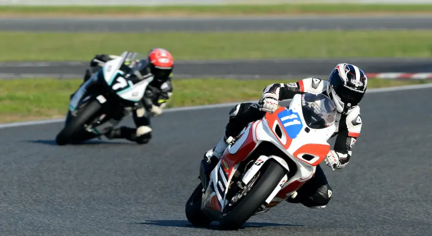 Dwóch motocyklistów w pełnym rynsztunku ściga się na torze. Jeden z nich, z numerem 11, pochyla się nisko w zakręcie, demonstrując emocje sportu motocyklowego.