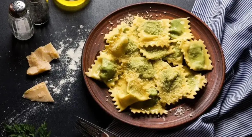 Pyszne ravioli z zielonym farszem, posypane parmezanem. Obok kawałki sera i przyprawy, gotowe do podania.