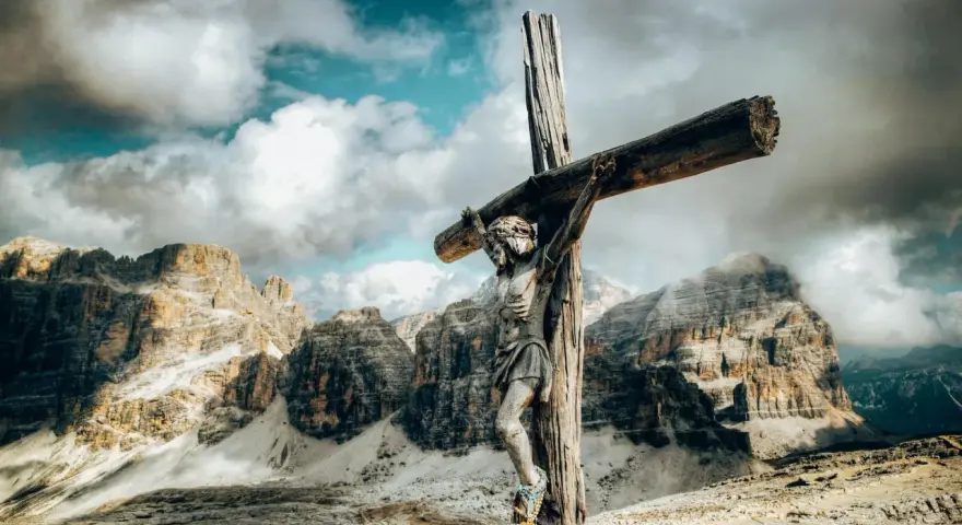Jesus am Kreuz, eine hölzerne Figur, steht vor majestätischen Bergen unter einem dramatischen Himmel.