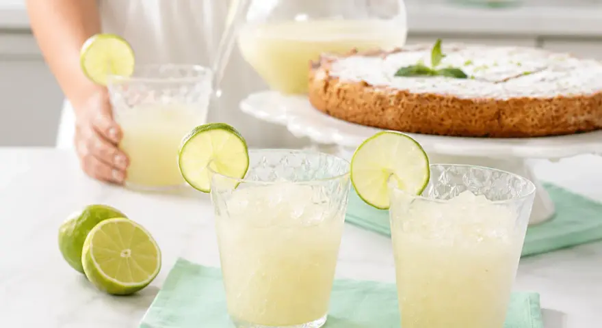 Dos vasos de granizado de limón refrescante, adornados con rodajas de lima, junto a limones frescos y un pastel.