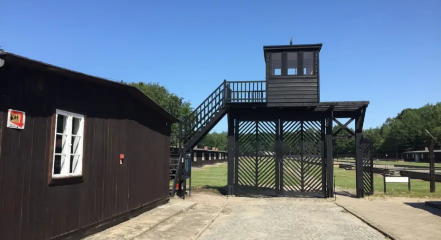 Brama i wieża strażnicza na terenie Muzeum Stutthof. Drewniane budynki i brukowany chodnik.