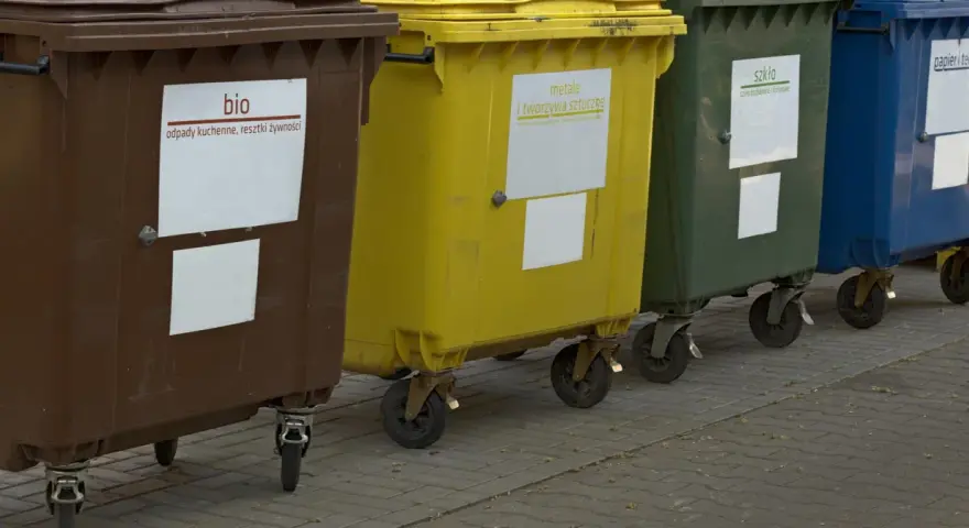Rząd kolorowych pojemników na śmieci: brązowy (bio), żółty (metale i tworzywa sztuczne), zielony (szkło), niebieski (papier i tektura).