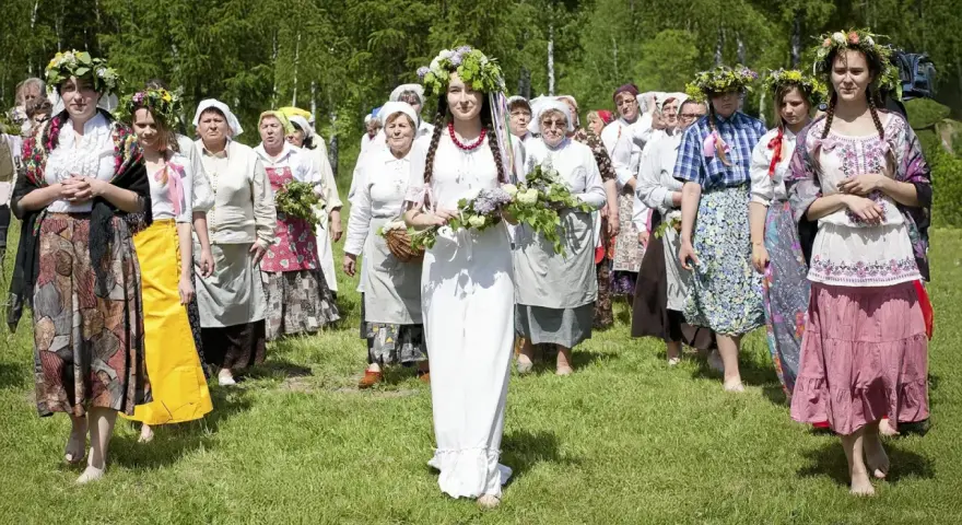 Grupa kobiet wiankach na głowach i tradycyjnych strojach idzie przez łąkę. Obraz nawiązuje do święta zielone świątki.