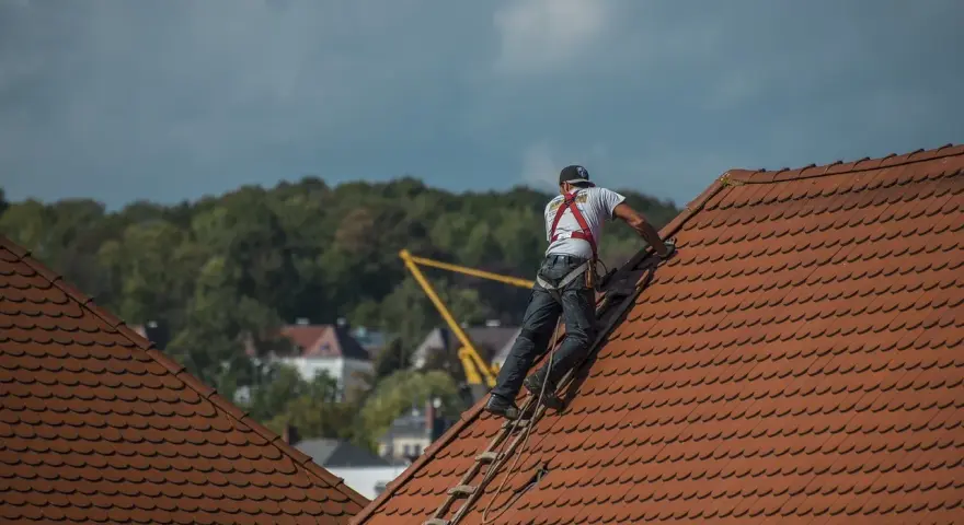 Robotnik w kasku i szelkach bezpieczeństwa na dachu, pracujący przy dachówce.