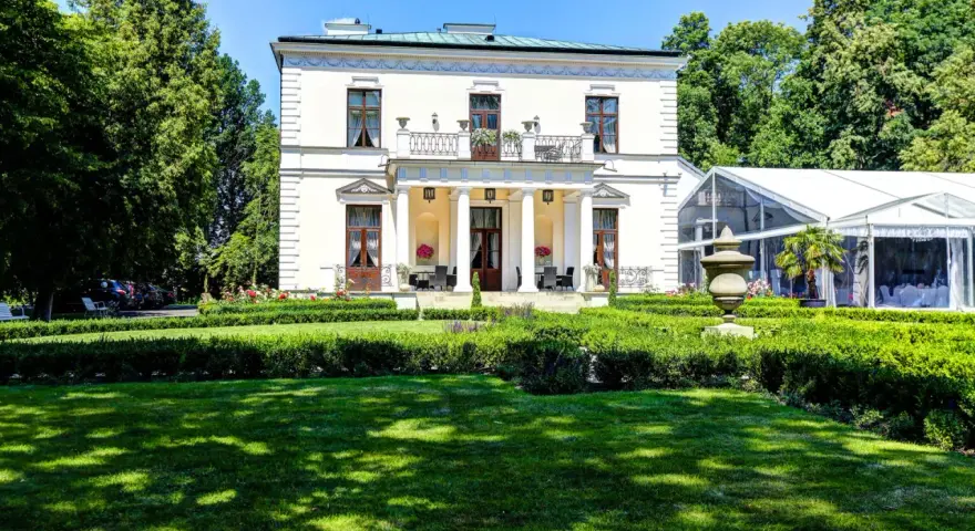Elegancki pałac Rozalin z kolumnowym portykiem i balkonem, otoczony zielonym ogrodem i nowoczesnym namiotem bankietowym.