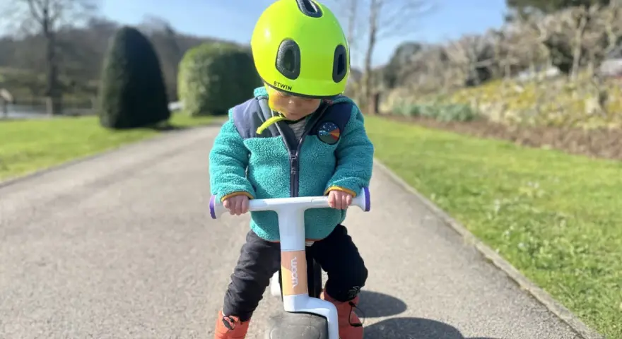 A child in a bright green helmet and teal jacket rides a woom wow balance bike on a sunny path.