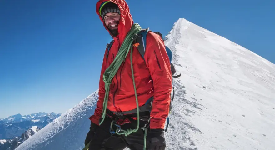 Uśmiechnięty alpinista na szczycie Mont Blanc, w czerwonej kurtce i z liną, gotowy na dalszą wspinaczkę.