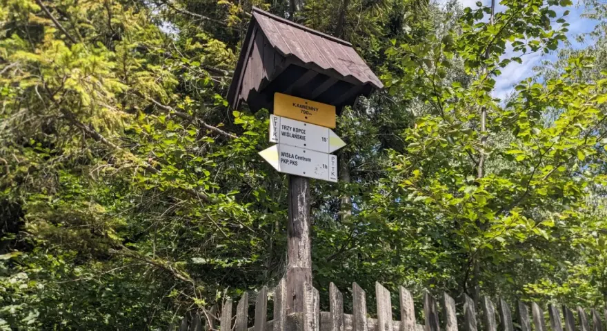 Znak szlaku PTTK w lesie, wskazujący drogę na Trzy Kopce Wiślańskie i do Wisły. W tle zieleń drzew i drewniany płot.