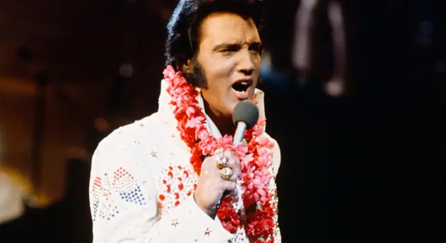 Elvis Presley singt in Hawaii, trägt einen weißen Anzug mit Blumenkette und Mikrofon.