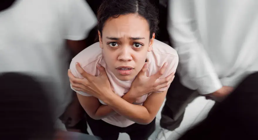 Donna spaventata, circondata da figure sfocate, con le braccia incrociate sul petto. Potrebbe rappresentare i disturbi psicosomatici, un elenco di sintomi fisici legati allo stress.