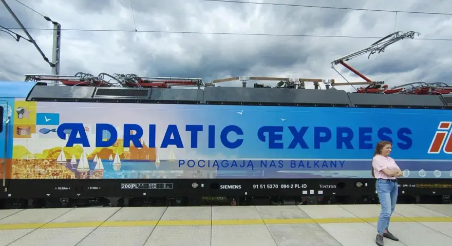 Kobieta stoi obok lokomotywy Adriatic Express z widokiem na morze i żaglówki.