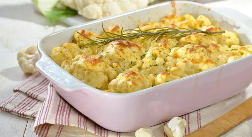 Pyszna keto zapiekanka z kalafiora w różowej naczyniu, udekorowana rozmarynem. Obok świeże różyczki kalafiora.