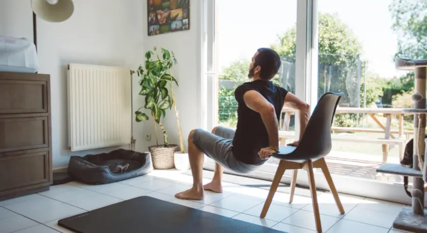 Un homme fait des dips sur une chaise, une idée de quel sport faire à la maison. Un tapis de yoga est au sol.