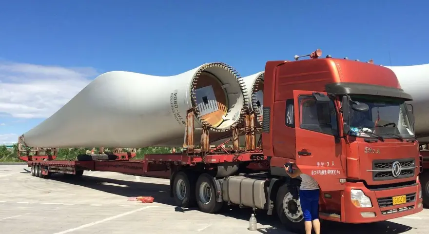 Pomarańczowy samochód ciężarowy przewozi ogromne łopaty turbin wiatrowych.
