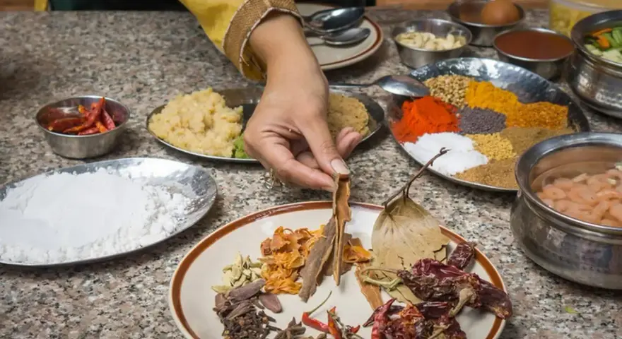 Ręka wybiera cynamon z talerza przypraw, przygotowując autentyczną kuchnię nepalską. Widać chili, mąkę, pastę imbirową i kolorowe przyprawy.