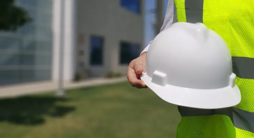Biały kask budowlany w dłoni, obok jaskrawożółtej kamizelki. Różne kolory kasków na budowie to symbol bezpieczeństwa.