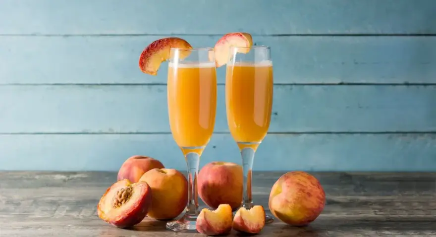 Dwa kieliszki z orzeźwiającym drinkiem na bazie likieru brzoskwiniowego, ozdobione plasterkami brzoskwini. Obok świeże owoce.