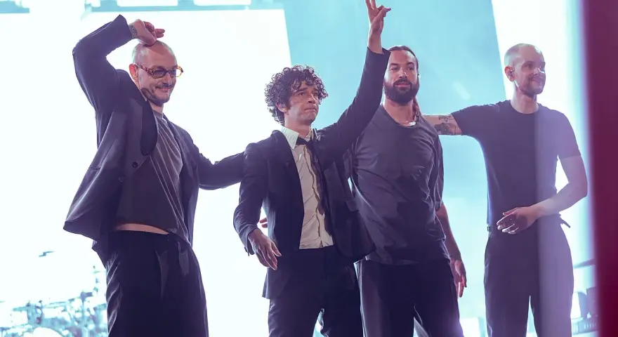 Matty Healy i zespół The 1975 na scenie. Muzycy pozdrawiają publiczność, jeden z nich unosi rękę w geście pokoju.