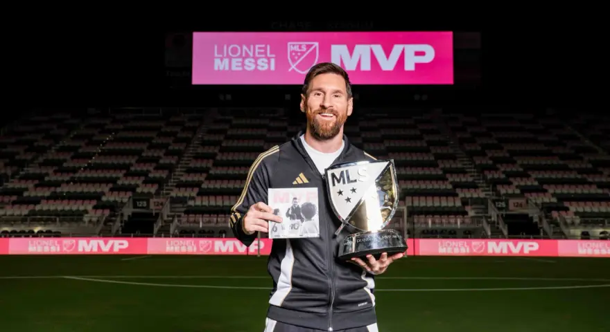 Lionel Messi sonríe en un estadio, sosteniendo un trofeo MLS MVP y un libro. El letrero del estadio dice "LIONEL MESSI MVP".