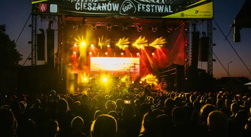 Tłumy bawią się na Cieszanów Rock Festiwal. Jasne światła sceny oświetlają zespół grający na tle zachodzącego słońca.