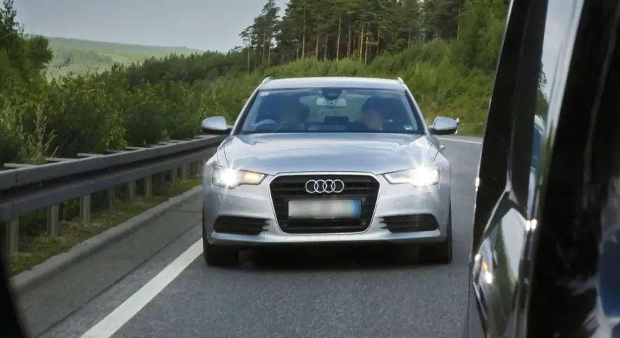 Silberner Audi auf der Autobahn, blendet mit Lichthupe. Vorbei an grünen Wäldern und einer Leitplanke.